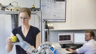 Eine wissenschaftliche Mitarbeiterin gibt einem bionischen Greifer einen Apfel